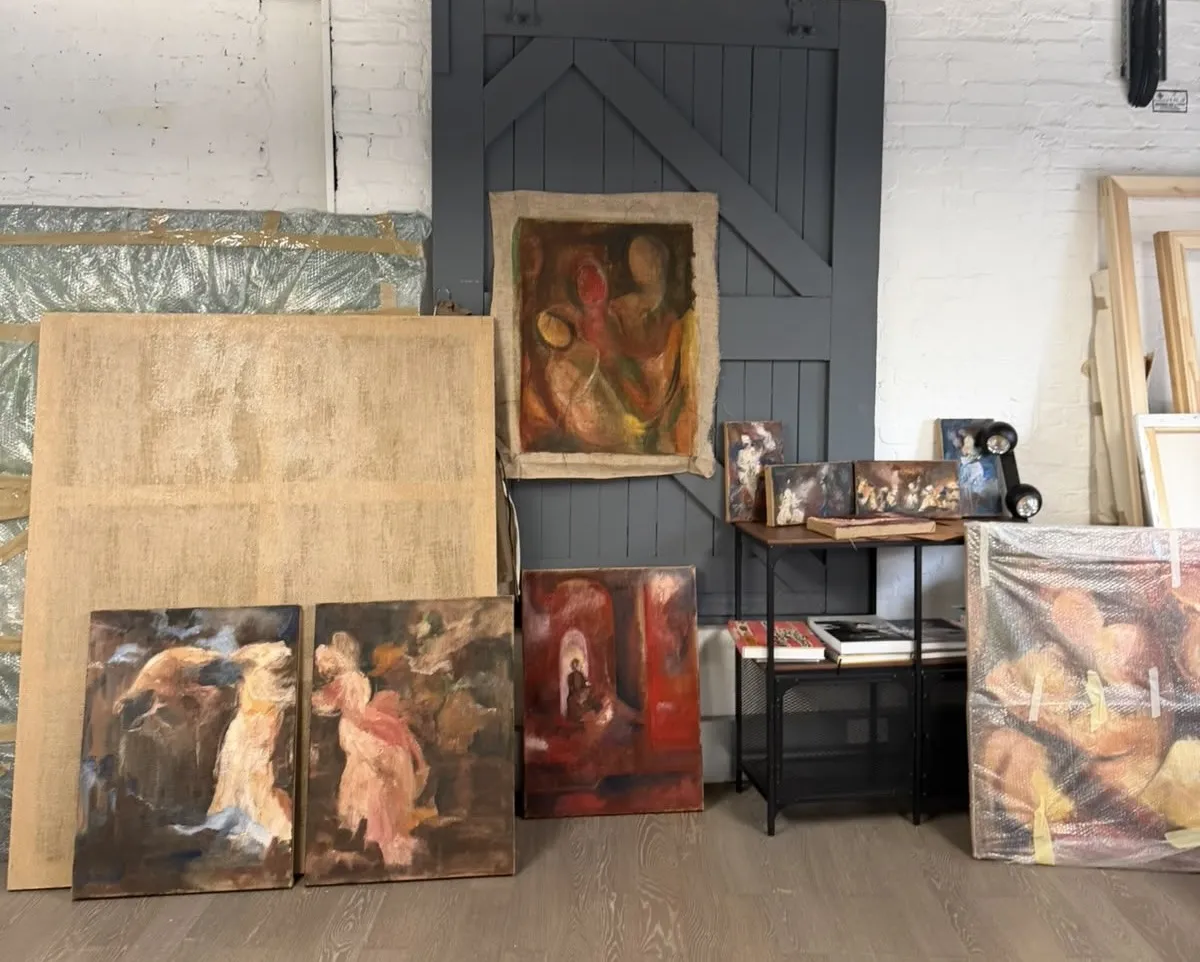A painter’s studio, with paintings on canvas and burlap leaning against a white brick wall and a grey wooden door